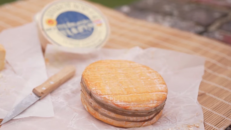 Un fromage Livarot rond à croûte orangée entouré de lanières de roseau posé sur du papier sulfurisé avec un couteau à côté